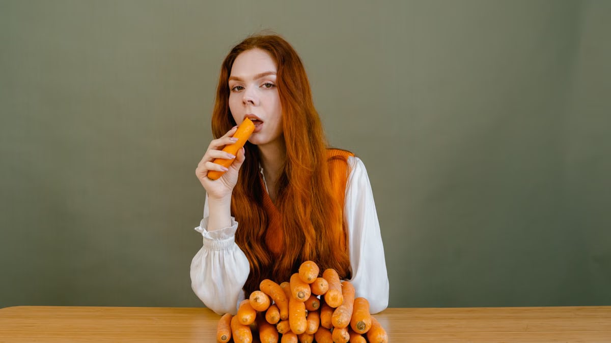 Red-haired woman sits at a table, holding a carrot, surrounded by a stack of vegetables.